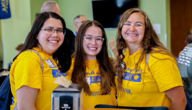 三名女学生穿着圣母大学同伴导师的黄色衬衫，在自助餐厅对着镜头微笑 